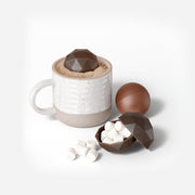 Zoku | Bomb Chocolate Mold: A ceramic mug with hot cocoa and a faceted chocolate bomb, with two chocolate spheres, one revealing marshmallows, nearby.