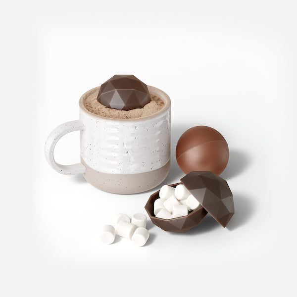 Zoku | Bomb Chocolate Mold: A ceramic mug with hot cocoa and a faceted chocolate bomb, with two chocolate spheres, one revealing marshmallows, nearby.