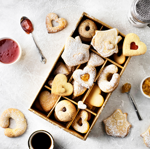 Austrian Holiday Cookies baking class product photo: A box of heart-shaped homemade cookies with jam and powdered sugar, surrounded by jam bowls, a shaker, and coffee.