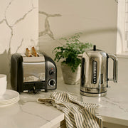 Dualit | 2 Slice Classic Toaster, Matt Black, with toast popping up, on a marble countertop alongside white dishes, a striped towel, and a potted plant.