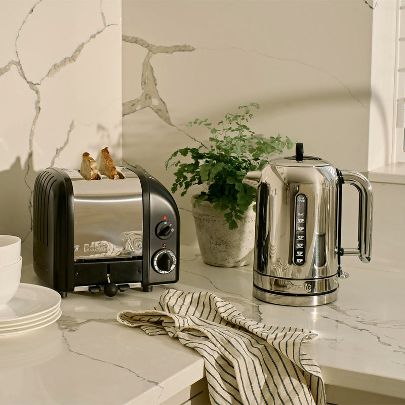 Dualit | 2 Slice Classic Toaster, Matt Black, with toast popping up, on a marble countertop alongside white dishes, a striped towel, and a potted plant.