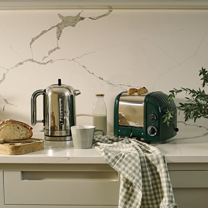 Dualit | 2 Slice Classic Toaster, Evergreen, showcased on a kitchen counter with bread, kettle, mug, milk bottle, and tea towel, highlighting its craftsmanship and functionality.