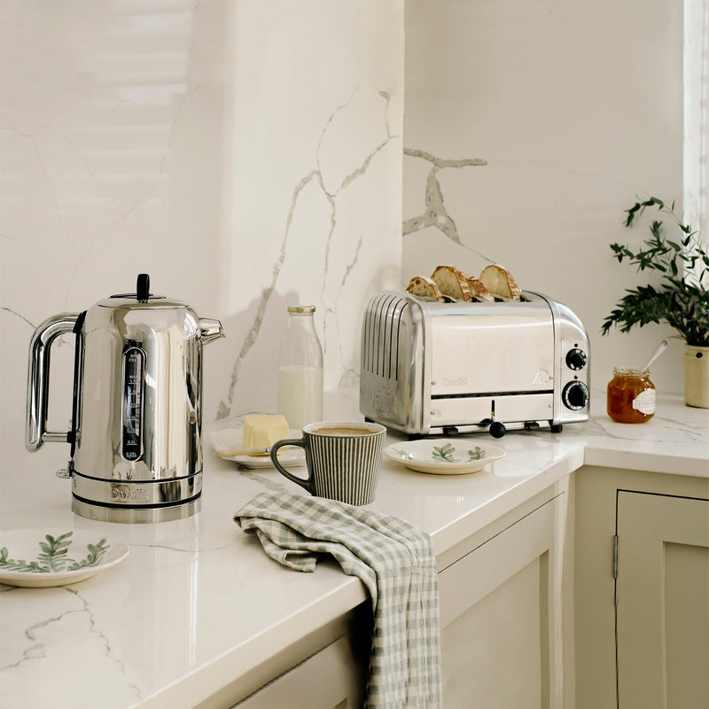 Dualit 4 Slice Classic Toaster, Polished, with toast popping up, showcased on a marble countertop set for breakfast alongside a kettle, mug, and jam.
