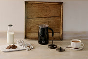 Dualit Hot Chocolate Maker and Milk Frother with whisk, lid, glass milk bottle, chocolate pieces, and a foamy drink in a cup on a light wood surface.
