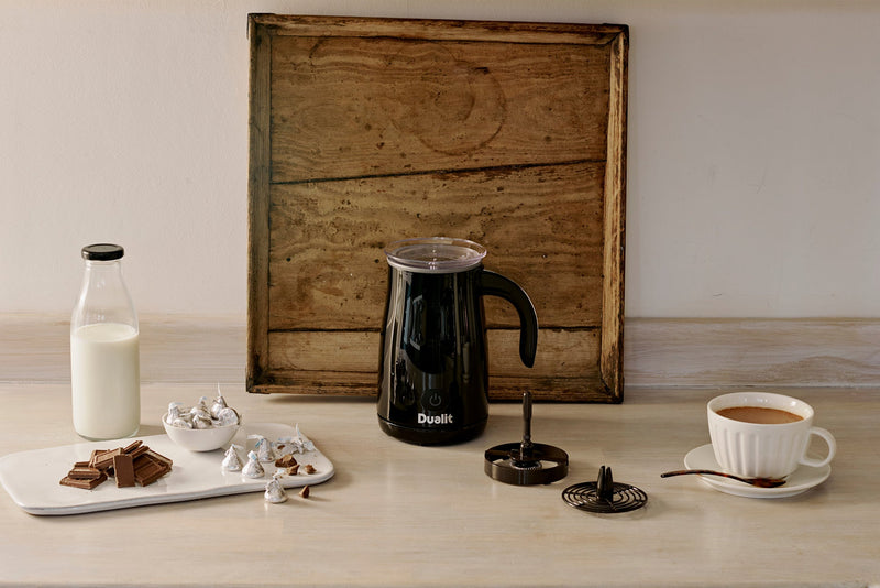 Dualit Hot Chocolate Maker and Milk Frother with whisk, lid, glass milk bottle, chocolate pieces, and a foamy drink in a cup on a light wood surface.