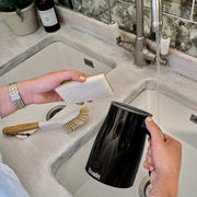 Hands wash a Dualit Hot Chocolate Maker and Milk Frother under a faucet in a kitchen sink, surrounded by cleaning tools and a potted plant.
