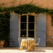 Outdoor setting with a Caravan Home 71 x 71 Forest Harvest Grey & Mustard Tablecloth on a table, showcasing hand-printed foliage and fruit design.