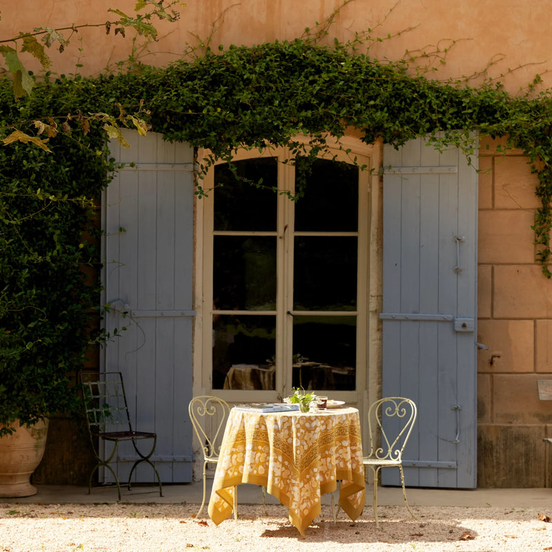 Outdoor setting with a Caravan Home 71 x 71 Forest Harvest Grey & Mustard Tablecloth on a table, showcasing hand-printed foliage and fruit design.