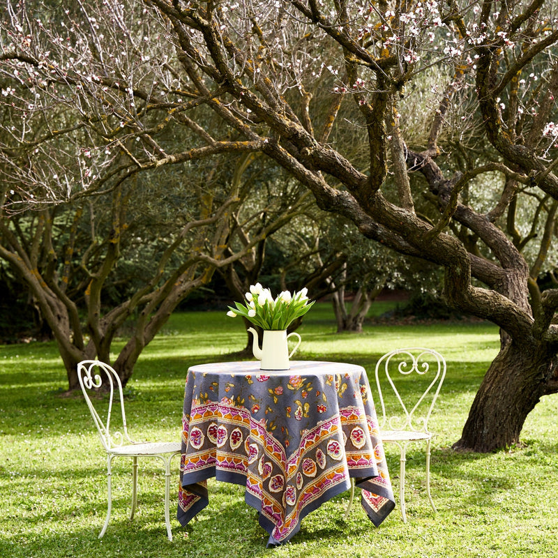 Pansy Red & Grey Tablecloth on outdoor table with white chairs under a tree, featuring floral patterns, handprinted craftsmanship from Caravan Home, enhancing a garden dining setting.