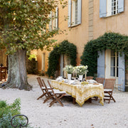 Caravan Home | 71 x 71 Forest Harvest Grey & Mustard Tablecloth elegantly drapes an outdoor table, showcasing hand-printed fruit and foliage motifs.