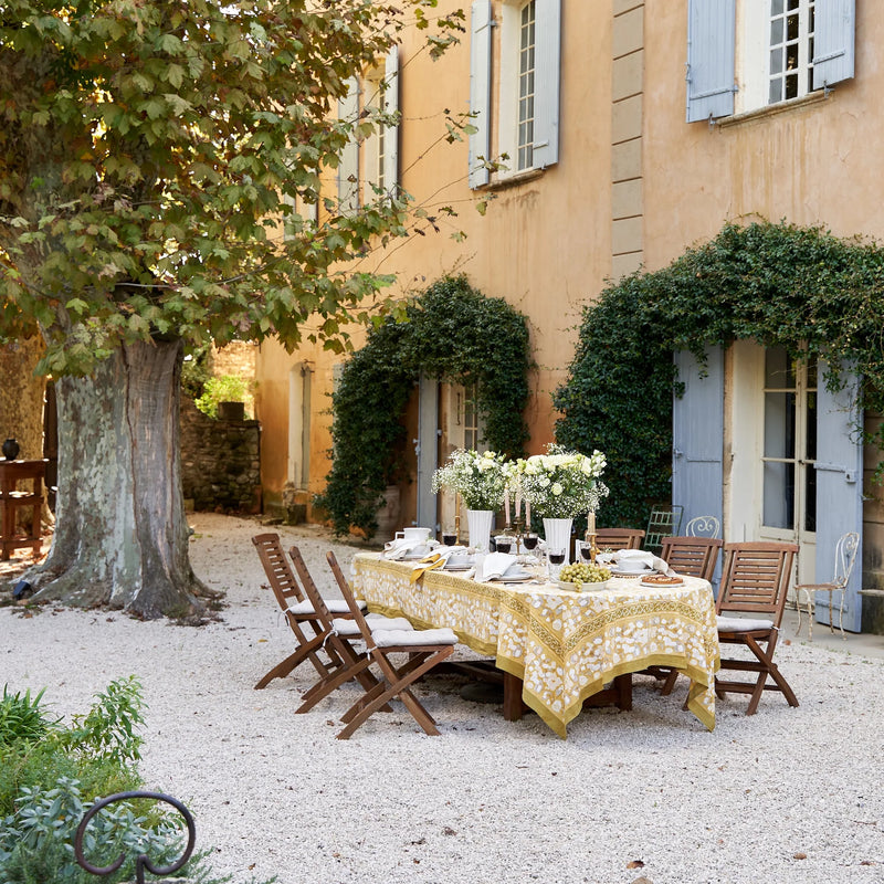 Caravan Home | 71 x 71 Forest Harvest Grey & Mustard Tablecloth elegantly drapes an outdoor table, showcasing hand-printed fruit and foliage motifs.