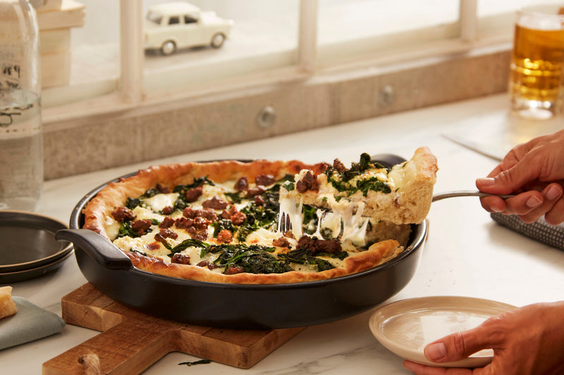 A slice of deep-dish pizza being lifted from the Emile Henry Chicago Deep Dish Pan, Charcoal, showcasing its crispy crust and melted cheese.