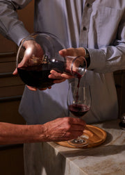 Holcomb Studio | Birdie Wine Decanter: A person elegantly pouring wine into a glass, showcasing the decanter's aerating sphere and minimalist ash wood trivet base.