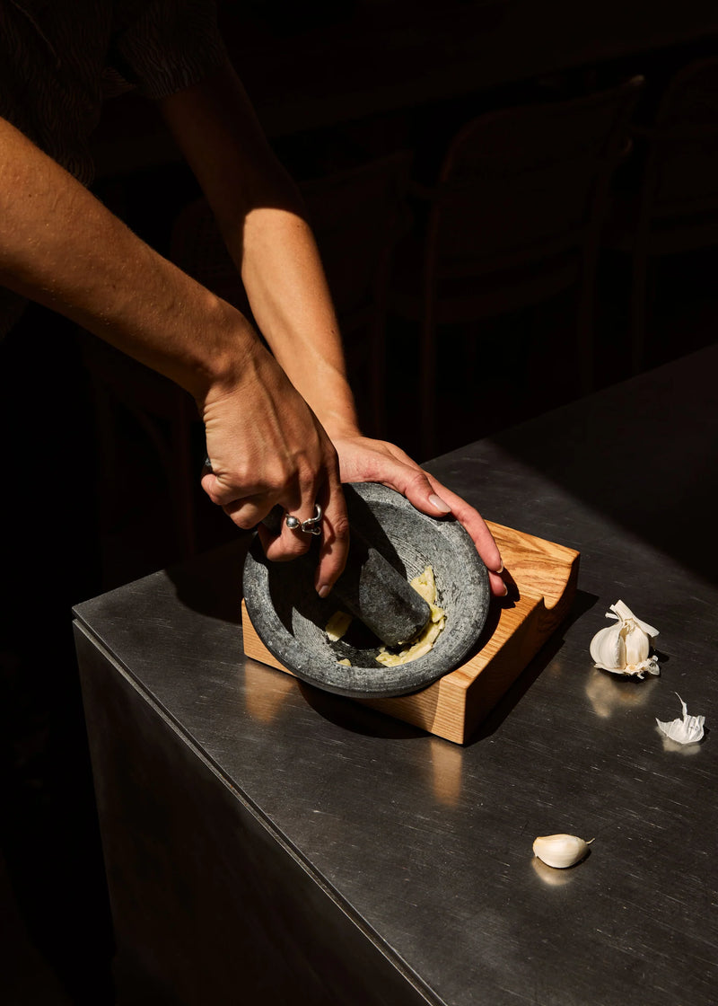 Holcomb Studio Perch Mortar & Pestle grinding garlic, showcasing its textured granite and ash wood base for enhanced kitchen functionality.