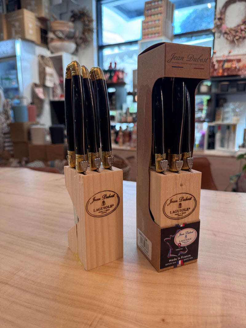 Jean Dubost | Laguiole Steak Knives w/Block (Set of 6), featuring black handles and brass-tone bolsters, displayed in a wooden block, partially encased in branded packaging.