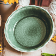 Person holding Emile Henry 175th Anniversary Pie Dish, Green/Ivory, over a wooden table with scattered yellow flower petals, celebrating French ceramic craftsmanship.