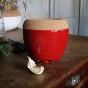 Emile Henry | Garlic Pot, Burgundy, a red ceramic pot with a cork lid, sits on wood beside a peeled garlic clove, ideal for storing garlic.