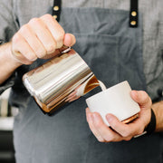 Barista pouring milk from FELLOW | Eddy Steaming Pitcher, 18 oz., into a cup, showcasing the precise design for crafting latte art.