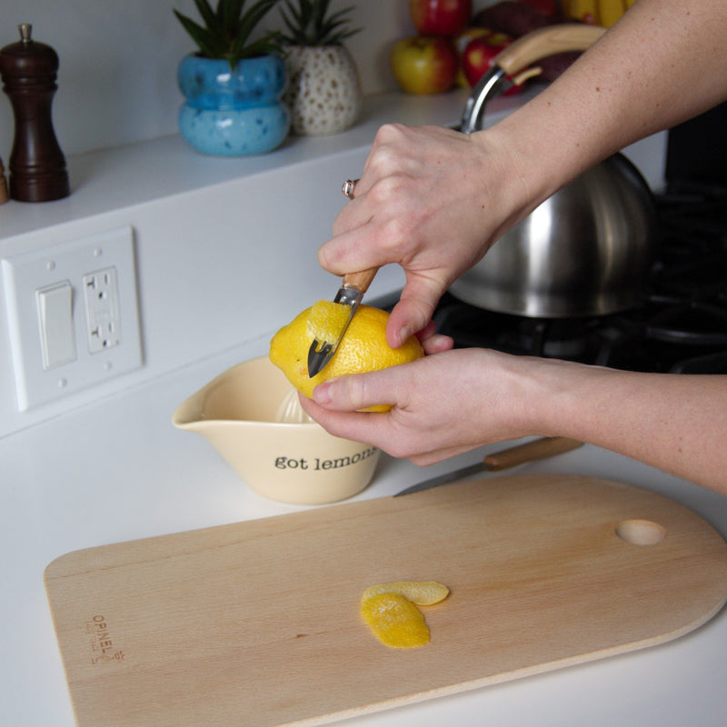 Person peeling lemon zest with Opinel paring knife from the Essential Small Kitchen Knife Set on a wooden board, featuring a ceramic bowl saying got lemons?.