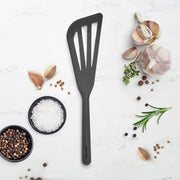 GreenPan slotted cooking turner on marble, surrounded by bowls of sea salt, peppercorns, garlic, coriander seeds, and herbs, reflecting Athens Cooks' curated kitchen tool selection.
