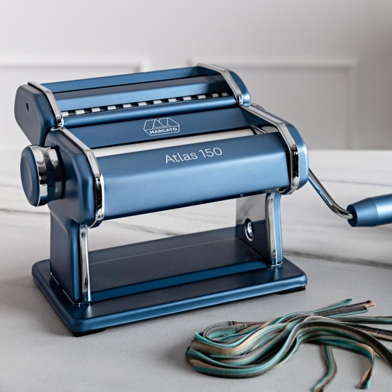 Marcato | Atlas 150 Pasta Maker (Ice) on a table, showcasing its manual handle and adjustable dial for rolling and cutting fresh pasta dough.