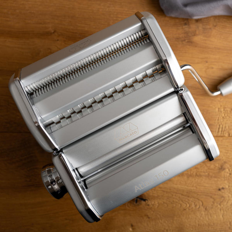 Marcato | Atlas 150 Pasta Maker on a table, showcasing its metal design and roller, ideal for crafting homemade lasagne, fettuccine, and tagliolini.