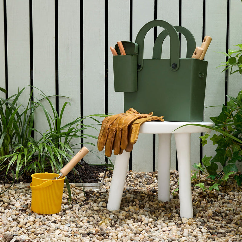 Hachiman Garden Tool Bag in garden setup, with tan leather gloves on a white stool, displaying its organized, easy-to-carry design with wooden-handled tools nearby.