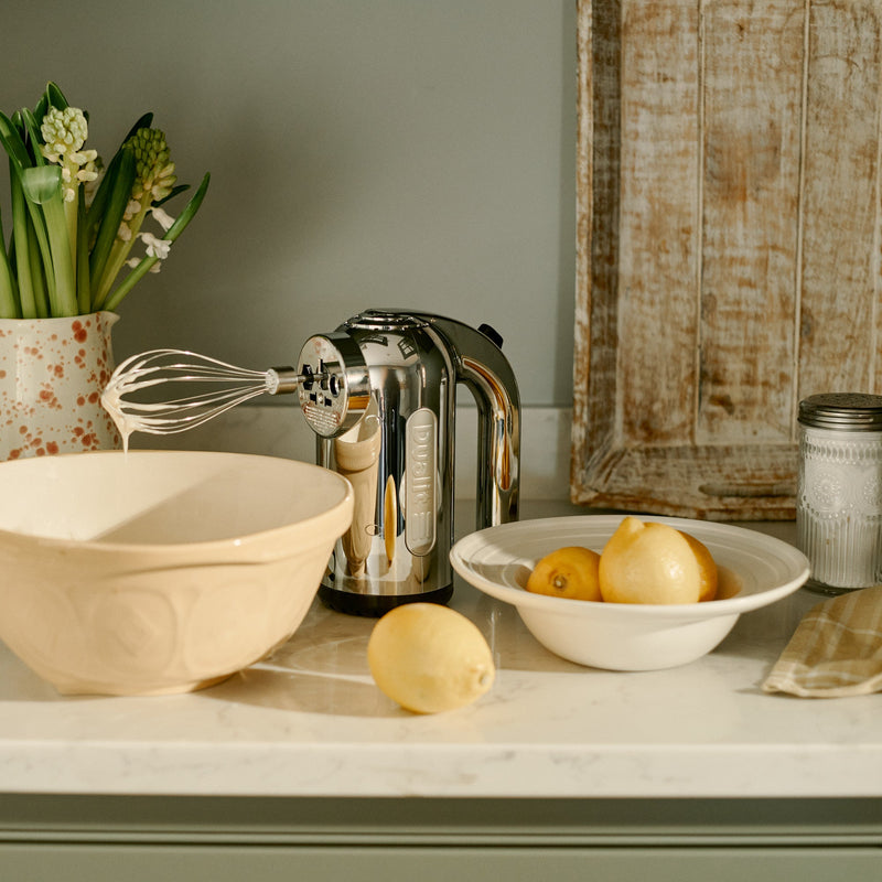 Dualit Hand Mixer in polished chrome with batter-covered whisk, set with a ceramic mixing bowl and lemons, suggesting a lemon baking project.