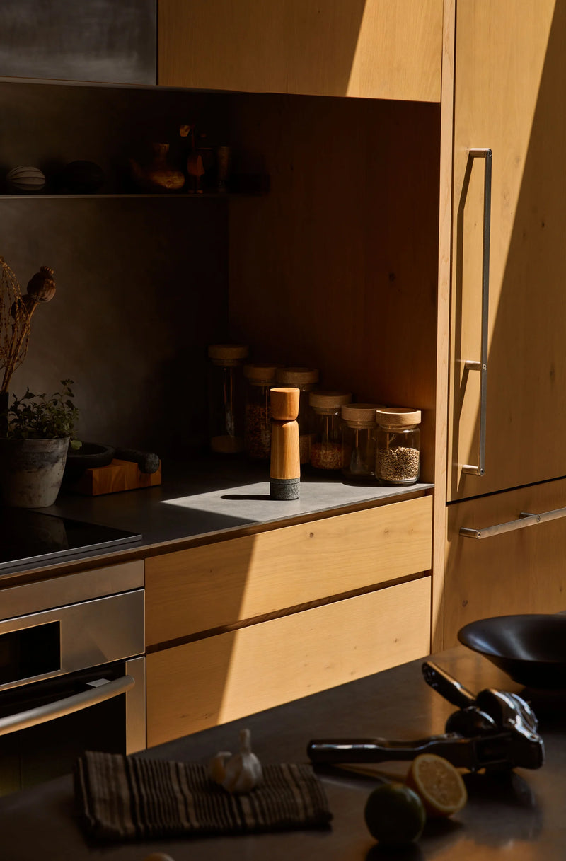 Holcomb Studio's Stone Pepper Mill on a wooden kitchen cabinet, surrounded by a salt shaker, jar of seeds, and a knife with garlic.