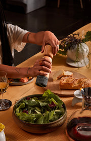 Holcomb Studio | Stone Pepper Mill in use, held over a dining table with salad, wine, and bread, highlighting its elegant design and functionality.
