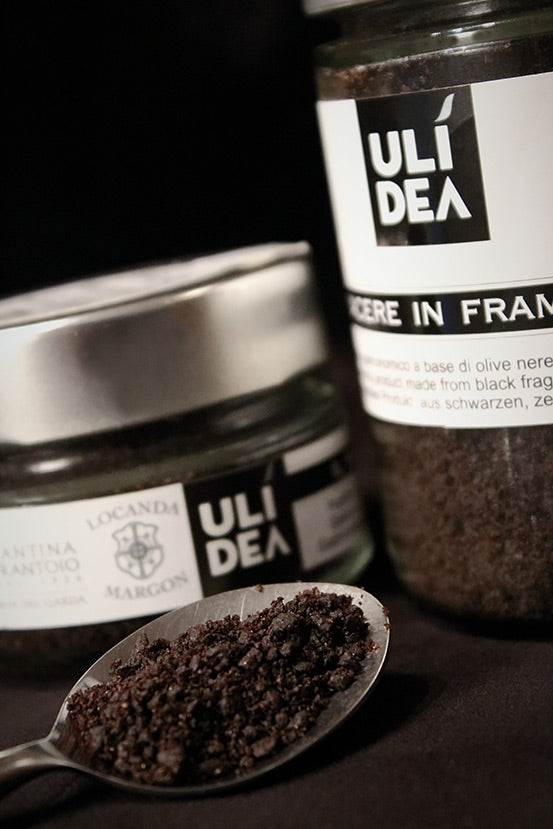 Ulidea | Black Olive Dust jar with a spoonful of the innovative topping beside it, crafted by Michelin Star Chef Ghezzi, ideal for enhancing culinary dishes.