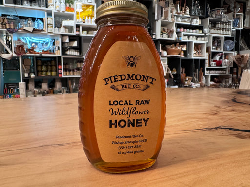 Piedmont Bee Company Local Raw Wildflower Honey jar on a wooden countertop, inside a specialty shop with shelves of kitchen goods in the background.