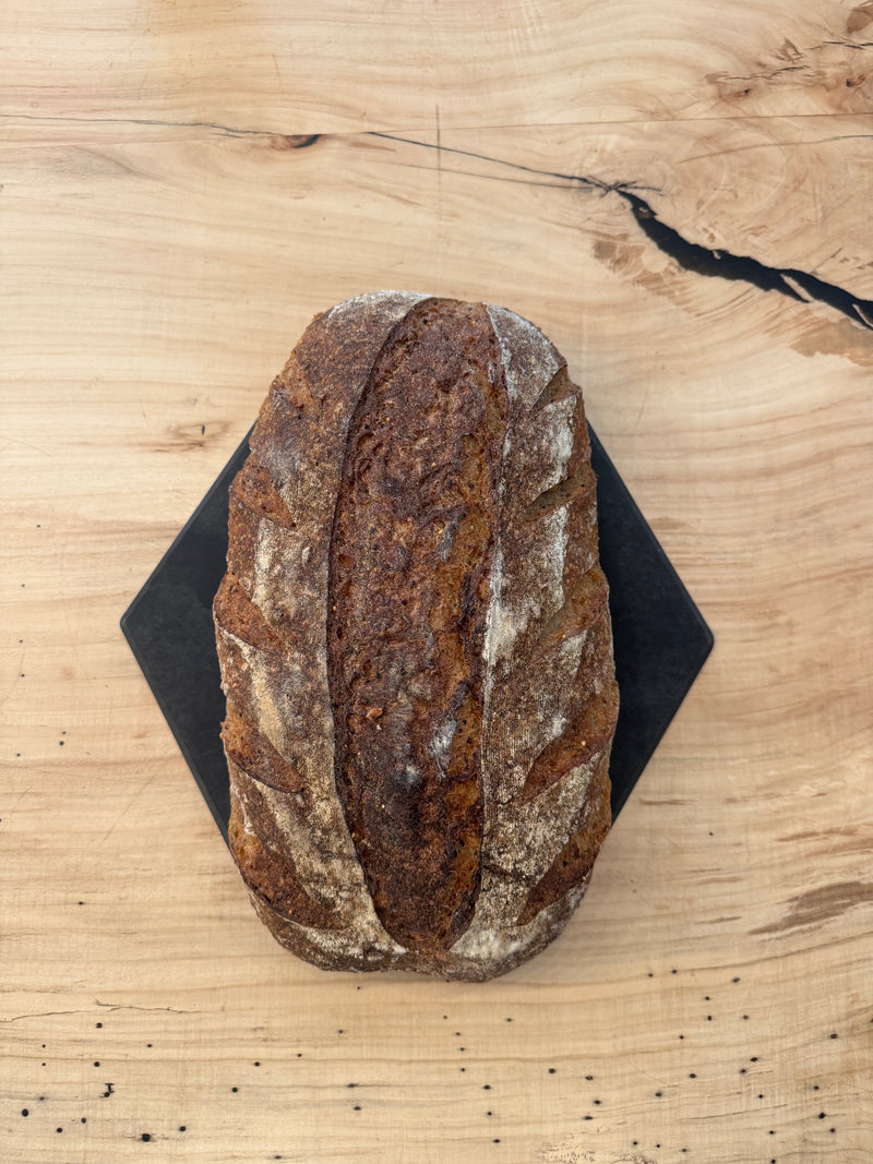 Independent Baking Co. | Multigrain Sourdough loaf with decorative scoring, on a dark cutting board, showcasing artisan craftsmanship ideal for everyday use.