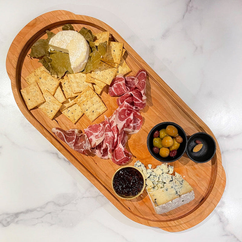 J.K. Adams Cherry Oval Wooden Serving Tray showcasing a charcuterie spread with cheeses, meats, crackers, olives, a nut, and jam, ideal for entertaining.