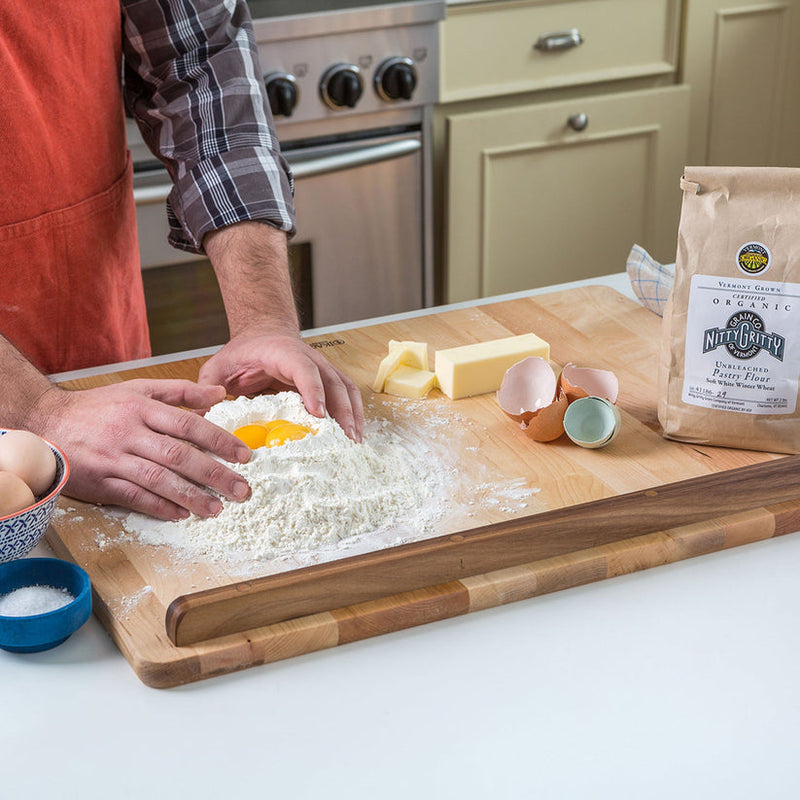 Person mixing flour and eggs on J.K. Adams | The Ultimate Pastry Board, featuring measurement guides and a cleat, surrounded by baking ingredients in a kitchen setting.