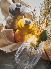 Peugeot | Bistro Salt Mill, Saffron, alongside a charcoal grinder, with a glass vase of dried grasses on a beige linen cloth in soft sunlight.