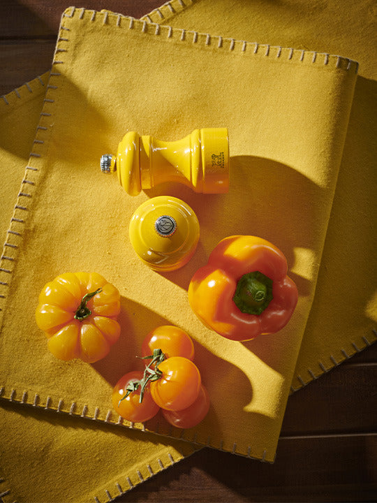 Peugeot | Bistro Pepper Mill, Saffron, disassembled on a mustard felt placemat with orange tomatoes and bell pepper, showcasing its vibrant design and culinary elegance.
