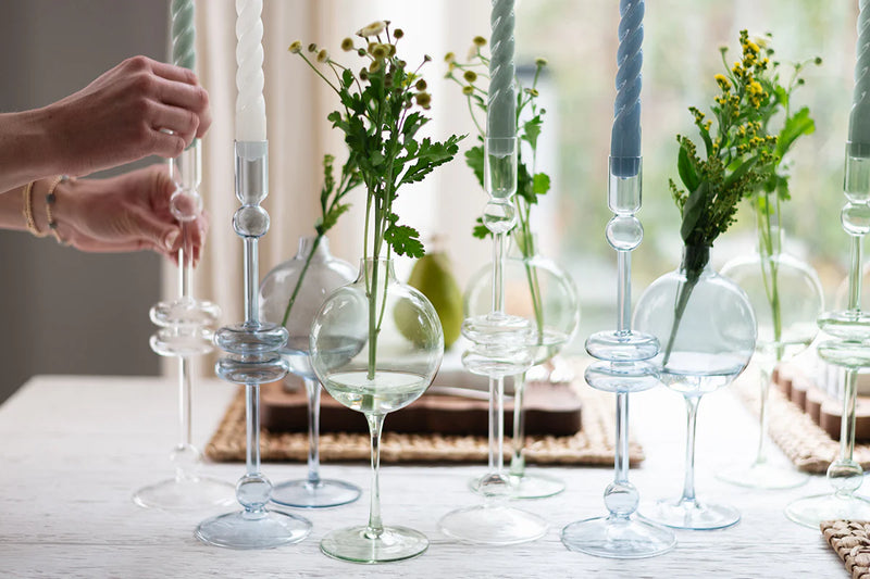 Clear Glass Candlestick Pair, Large by etú HOME, elegantly displayed among glass vases with flowers, capturing a sophisticated table setting.