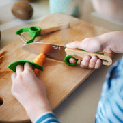 Child hands using Opinel | Le Petit Chef 3pc Knife Set - Green to safely slice a carrot, featuring a knife, finger guard, and peeler for young chefs.
