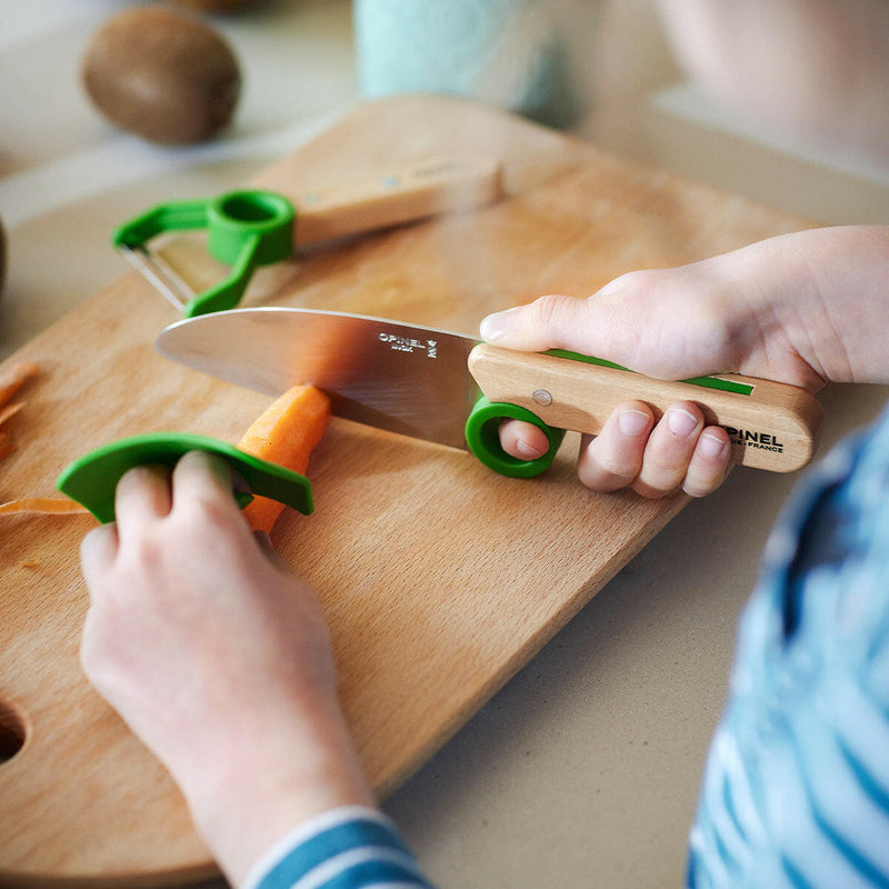 Child hands using Opinel | Le Petit Chef 3pc Knife Set - Green to safely slice a carrot, featuring a knife, finger guard, and peeler for young chefs.
