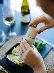 Peugeot | Line Pepper Mill in use over a cheese-topped dish, set beside a salad and wine, reflecting high-quality French craftsmanship.