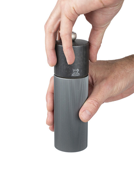 Hands holding a Peugeot | Line Pepper Mill, Aluminum + Beechwood, Carbon Finish, 7 inches, showcasing adjustable grind knob and Peugeot logo, reflecting French craftsmanship.