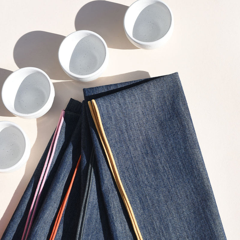 Atelier Saucier | Rainbow Denim Napkins, Set of 4, featuring colorful edges, stacked beneath four white ceramic bowls, perfect for adding fun to your dining experience.
