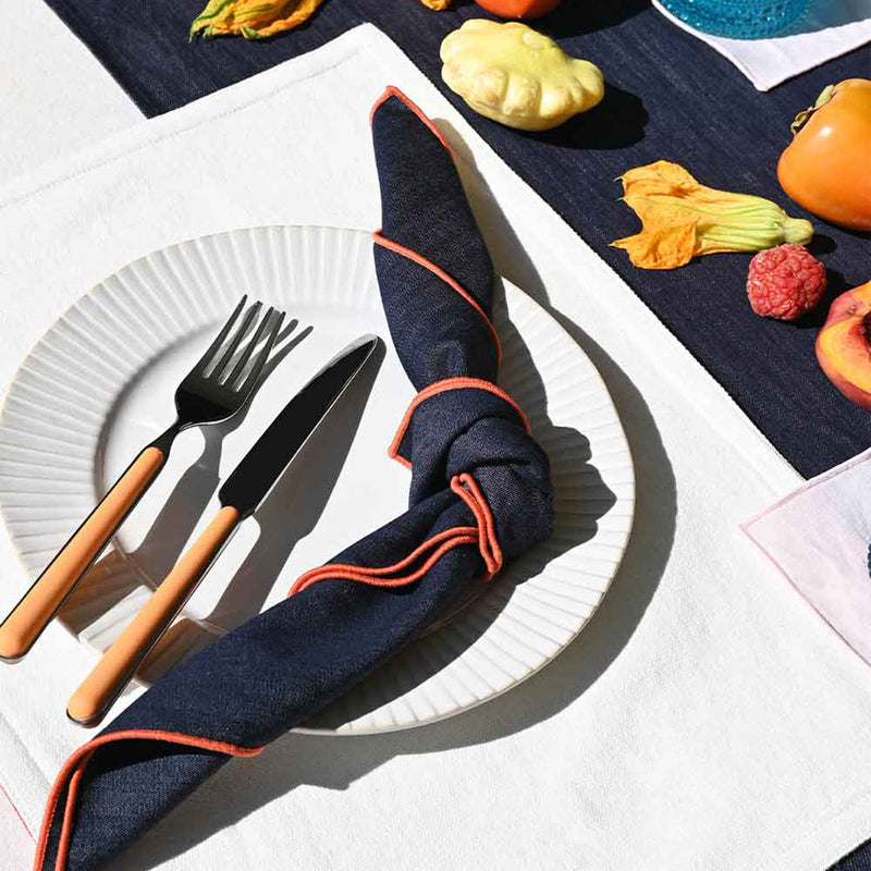 Atelier Saucier Rainbow Denim Napkin, edged in bright colors, is knotted on a fluted white plate with peach-handled cutlery, complemented by autumnal garnishes.