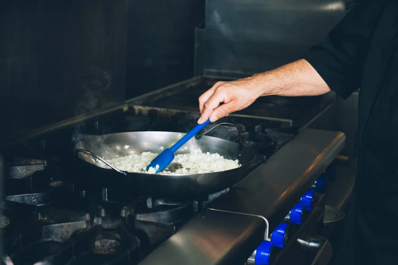 A person stirs onions in a Matfer Bourgeat Black Carbon Steel Paella Pan on a professional stovetop, showcasing its durability and heat retention.