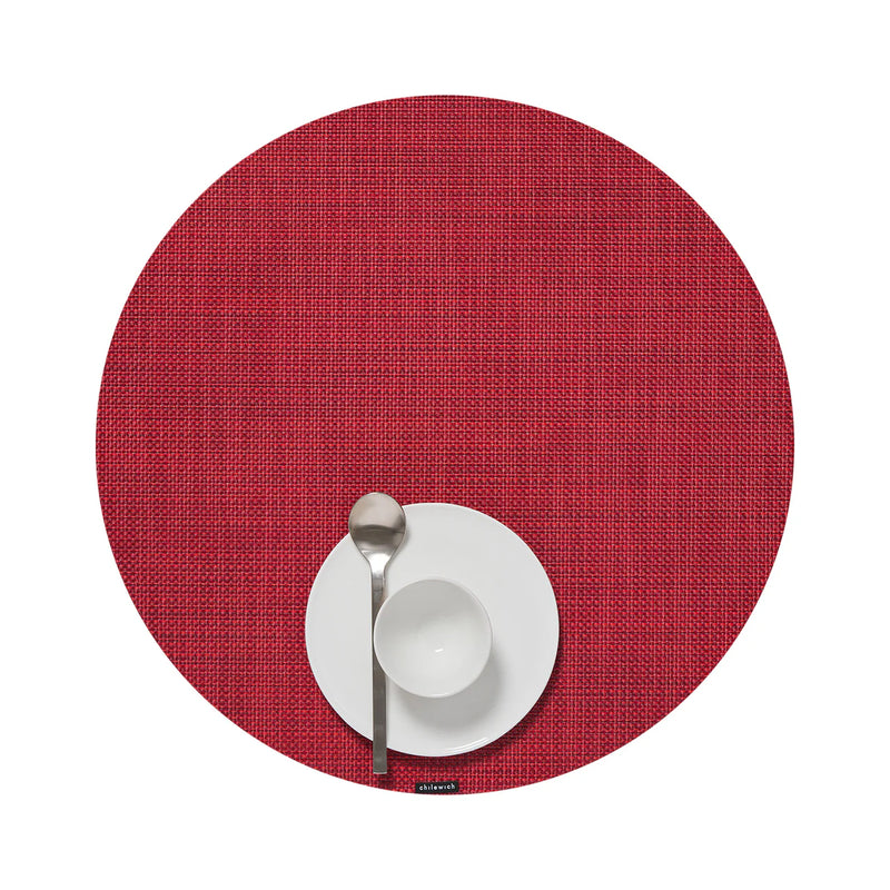 Chilewich | Mini Basketweave Round Placemat, Scarlet, displayed with a white dinner plate, bowl, and silver spoon, showcasing its textured design in a simple table setting.