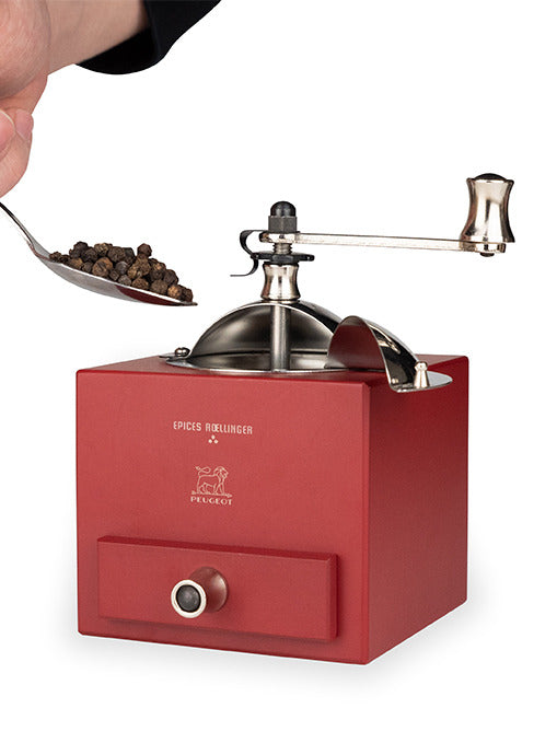 Peugeot | Roellinger Pepper Mill, Red: A red, box-style manual grinder being filled with black peppercorns, featuring a drawer for fresh pepper collection.