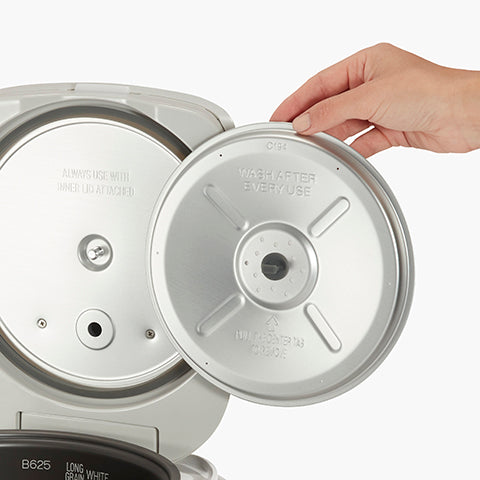 Hand lifting inner lid of Zojirushi | Micom Rice Cooker Warmer - Stainless Gray, highlighting metal insert with usage instructions, showcasing durable design for kitchen efficiency.