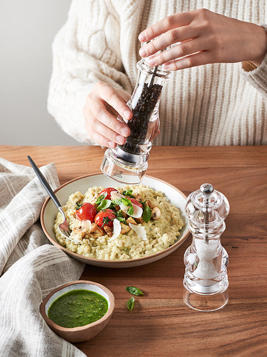 Hands seasoning risotto with the Peugeot Nancy 12-inch acrylic pepper mill, showcasing its clear design and adjustable coarseness beside a matching salt mill.