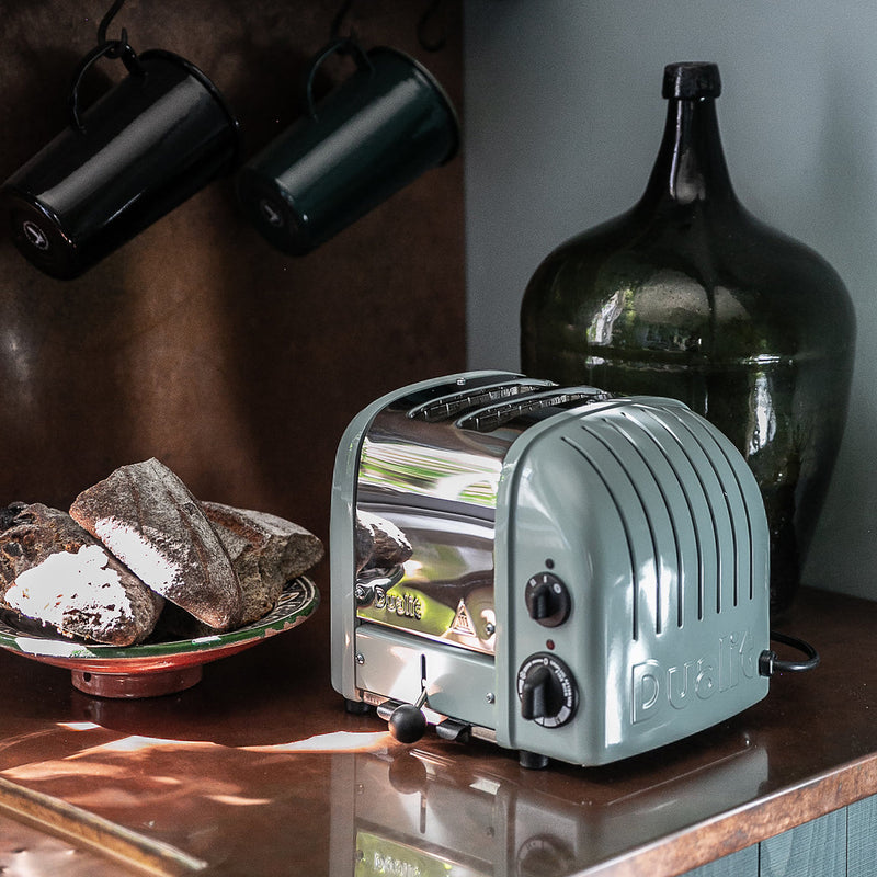 Dualit 2 Slice Classic Toaster, Eucalyptus, on a kitchen counter with rustic bread and vintage decor, showcasing its elegant design and craftsmanship.